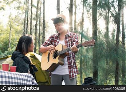 Portrait of Asian man wearing check shirt and hat playing guitar and singing for his beautiful girlfriend while camping in holidays. Travel and Summer concept.