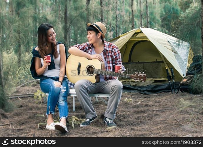 Portrait of Asian man wearing check shirt and hat playing guitar and celebrate to his beautiful girlfriend while camping in holidays. Travel and Summer concept.