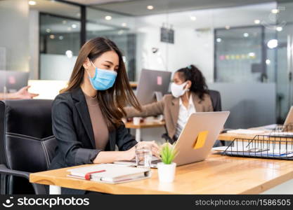 Portrait of asian office employee businesswoman wear protective face mask work in new normal office with interracial team in background as social distance practice prevent coronavirus COVID-19.