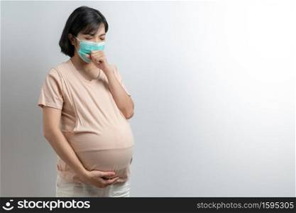Portrait of Asian Pregnant woman wearing protective face mask coughing on white isolated background. Young mother having a cold or flu standing touching her belly with copy space during covid pandemic