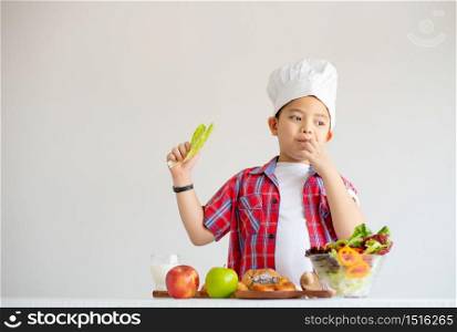 Portrait of Asian red shirt little boy show eat vegetable in room with white background and stand with various types of vegetables and fruits also milk.
