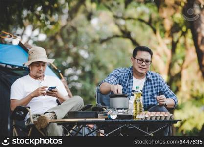 Portrait of Asian relaxed man using smartphone and waiting for his friend cooking with tent background in c&ing. Cooking set front ground. Outdoor cooking, traveling, c&ing, lifestyle concept.