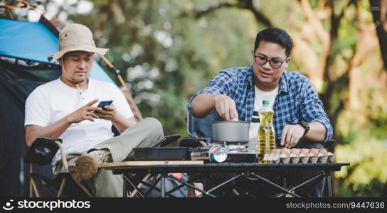 Portrait of Asian relaxed man using smartphone and waiting for his friend cooking with tent background in camping. Cooking set front ground. Outdoor cooking, traveling, camping, lifestyle concept.