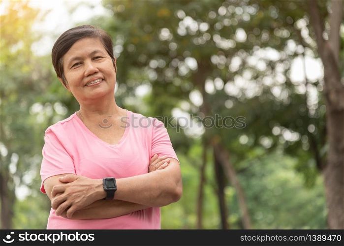 Portrait of Asian Senior woman in pink shirt smiling with arms crossed before exercising at park outdoor. Good health. Copy space.