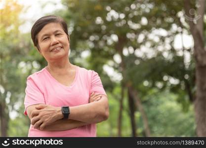 Portrait of Asian Senior woman in pink shirt smiling with arms crossed before exercising at park outdoor. Good health. Copy space.
