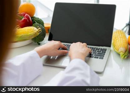 Portrait of Asian smiling female nutritionist typing on laptop computer for balanced corrective diet plan for patient or weight loss advice. Concept of healthy lifestyle, healthy food, dieting
