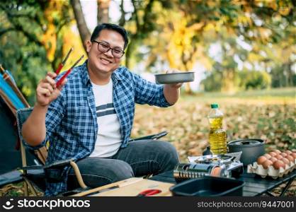 Portrait of Asian traveler man glasses frying a tasty fried egg in a hot pan at the campsite. Outdoor cooking, traveling, camping, lifestyle concept.