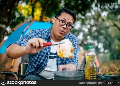 Portrait of Asian traveler man glasses pork steak frying, BBQ in roasting skillet pan or pot at a campsite. Outdoor cooking, traveling, camping, lifestyle concept.