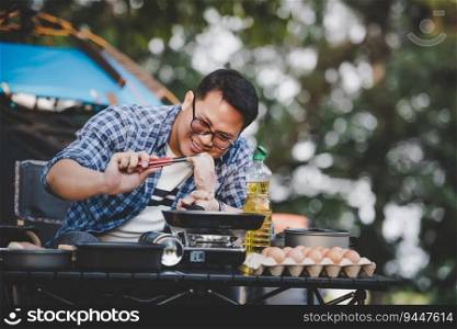 Portrait of Asian traveler man glasses pork steak frying, BBQ in roasting skillet pan or pot at a campsite. Outdoor cooking, traveling, camping, lifestyle concept.