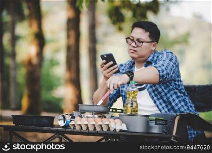 Portrait of Asian traveler man glasses using smartphone while cooking a meal at a campsite. Outdoor cooking, traveling, camping, lifestyle concept.
