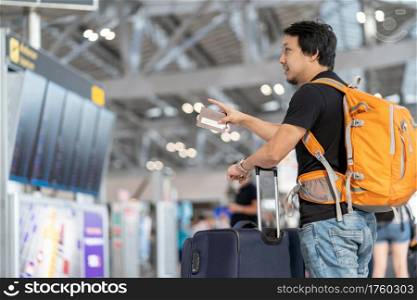 Portrait of Asian traveler with luggage with passport looking and pointing over the flight board for check-in at the flight information screen in modern an airport, travel and transportation concept.