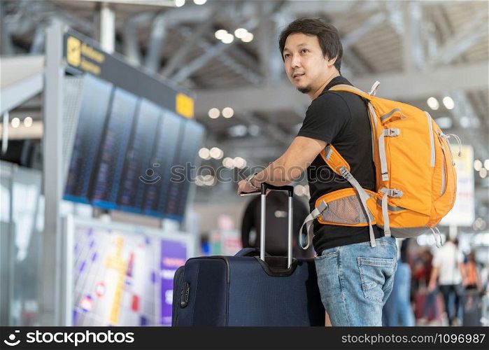 Portrait of Asian traveler with luggage with passport standing over the flight board for check-in at the flight information screen in modern an airport, travel and transportation with technology concept.