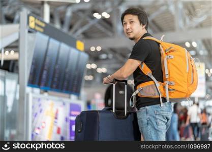 Portrait of Asian traveler with luggage with passport standing over the flight board for check-in at the flight information screen in modern an airport, travel and transportation with technology concept.