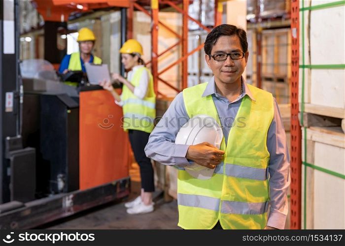 Portrait of Asian warehouse manager hold hard hat with warehouse worker operate forklift to check inventory in background. Reopening business warehouse technology and logistic concept.