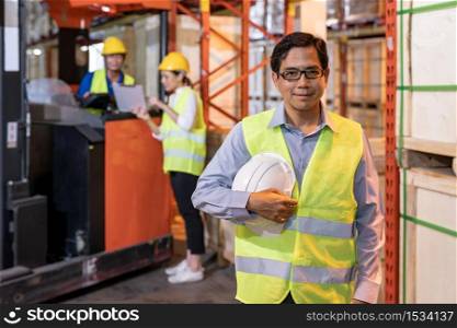 Portrait of Asian warehouse manager hold hard hat with warehouse worker operate forklift to check inventory in background. Reopening business warehouse technology and logistic concept.