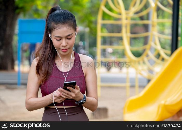 Portrait of Asian woman concentrate to use mobile phone and stay near playground during exercise in park or garden with morning light.