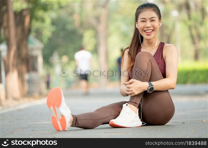 Portrait of Asian woman look at camera and sit with one&rsquo;s knees up on road of green park or garden with morning light.