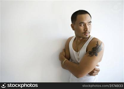 Portrait of Asian young adult man leaning against white wall with arms crossed looking over shoulder.