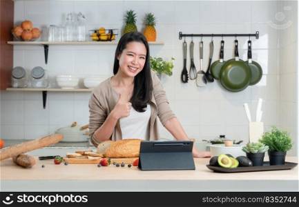 Portrait of Asian young beautiful woman standing and thumbs up in the kitchen and cooking healthy food with bread, fruits and vegetables. Smiling while using tablet computer