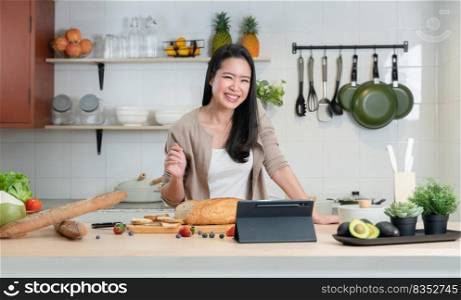 Portrait of Asian young beautiful woman standing in the kitchen and cooking healthy food with bread, fruits and vegetables. Smiling and watching tablet computer