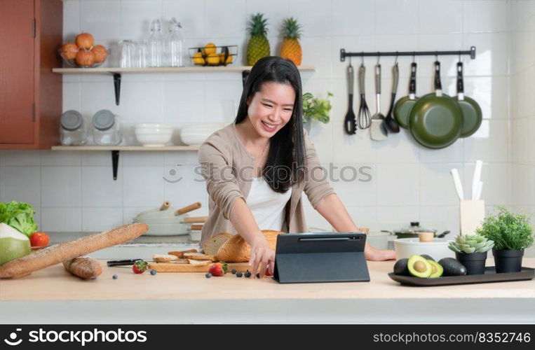 Portrait of Asian young beautiful woman standing in the kitchen and cooking healthy food with bread, fruits and vegetables. Smiling, watching and touching screen tablet computer