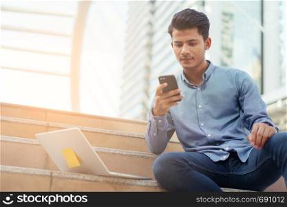 Portrait of asian young businessman sit staircase at Outside Office. Young businessman wear shirt smiling and looking at smartphone. Laptop