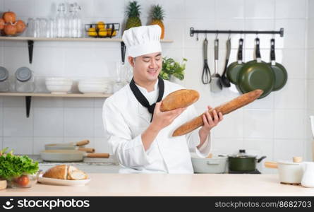 Portrait of Asian young handsome chef man wear white chef uniform and hat, holding loaf of handmade french bread in hands, smiling looking at breads, working at restaurant kitchen