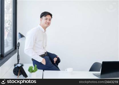 Portrait of Asian young smart handsome businessman wear white shirt holding formal suit in hand sitting on desk smiling at camera. Confident man working with laptop at office. White background