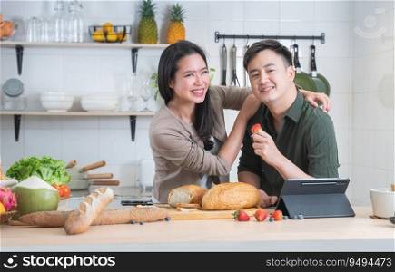 Portrait of Asian young sweet couple lover cooking breakfast together in home kitchen. Beautiful wife smiling embracing her husband while he eating strawberry and working on tablet. Looking at camera