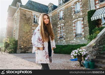 Portrait of atrrative woman wears white loose cape, stands outdoor against ancient builduing, has excursion in ancient city, has serious dreamful expression, looks directly in camera. Having nice day