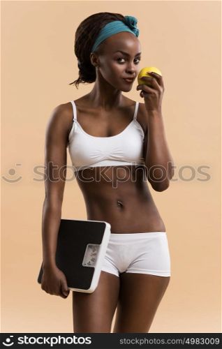Portrait of attractive african smiling woman eating green apple on beige studio background, looking at camera