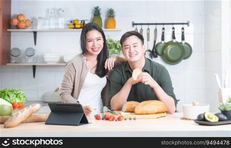 Portrait of attractive Asian young couple using tablet at kitchen, smiling, looking at camera, enjoy cooking preparing healthy breakfast meal together at home. Family lifestyle and food concept