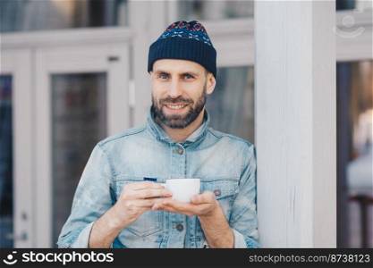 Portrait of attractive bearded male has pleasant smile, dressed in denim jacket, holds white cup of coffee or tea, enjoys good time, has coffee break. Happy handsome stylish young hipster guy