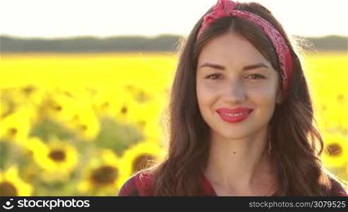 Portrait of attractive brunette female in red headband and lipstick with perfect teeth posing in nature, looking with sensual smile on blurry sunflower background. Pretty woman with brown eyes gazing at camera with radiant smile on summer day.