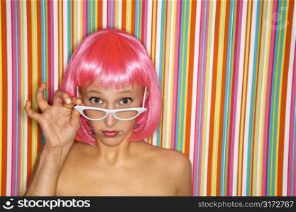 Portrait of attractive Caucasian young adult woman wearing pink wig against striped background peeking over glasses at viewer.
