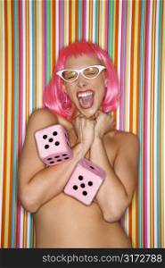 Portrait of attractive Caucasian young adult woman wearing pink wig against striped background holding large pink dice and making sassy expression.