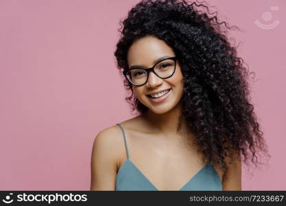 Portrait of attractive dark skinned woman with Afro bushy hair, dressed casually, looks through transparent glasses, has happy smile on face, isolated on pink background. Happy emotions concept
