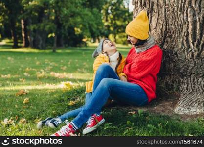Portrait of attractive female model, has pleasant conversation with daughter, rest on green grass, look happily, have joy. Lovely woman with small girl sit on ground, admire autumn and fresh air