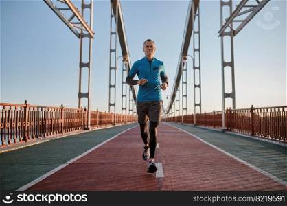 Portrait of attractive fit man running fast along big modern bridge road. Jogger doing outdoors workout in morning. Attractive fit man running fast along big modern bridge