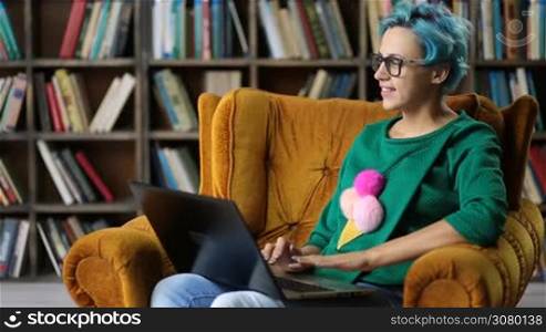 Portrait of attractive hipster student girl in eyeglasses pondering preparing report for homework with laptop pc as she sits in comfotable chair in library. Thoughtful gorgeous young woman dressed in stylish outfit concentrated on writing an article.