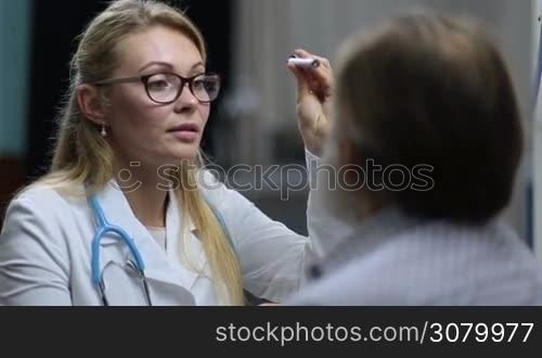 Portrait of attractive long blonde hair female physician checking pupillary reflex of senior male patient with flashlight in medical office. Attentive female neurologist testing reflexes of elderly man&acute;s eyes in clinic during medical checkup.