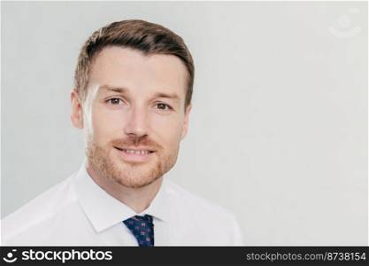 Portrait of attractive male CEO with confident facial expression, looks confidently directly at camera, wears elegant luxury clothes, poses against white studio wall with blank space on left for text