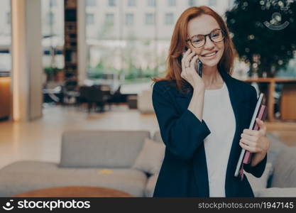 Portrait of attractive positive european business woman in elegant stylish outfit having mobile conversation, making business call, talking on smartphone while waiting for client or partner indoors. Portrait of attractive positive european business woman having mobile conversation