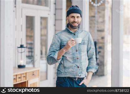 Portrait of attractive stylish bearded male with happy look, wears fashionable denim jacket, hat and trousers, holds mug of hot beverage, recreats at home after hard working day, smiles pleasantly