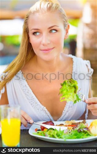Portrait of attractive woman eating fresh green salad using knife and fork, having lunch in outdoors restaurant, healthy lifestyle concept