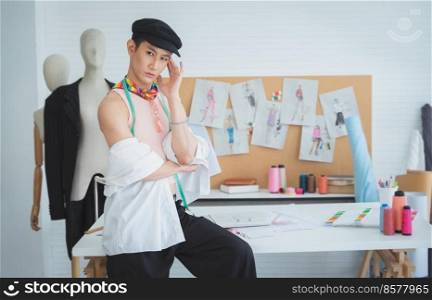 Portrait of attractive young Asian LGBTQ gay dressmaker sitting pose with tape measure on working desk, confident looking at camera, sketch of fashion dress design, equipments and mannequin in studio