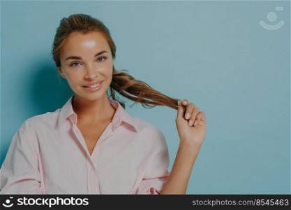 Portrait of attractive young female model with combed hair holds pony tail smiles gently looks directly at camera dressed in formal shirt isolated over blue background copy space for your advert. Portrait of attractive young female model with combed hair holds pony tail smiles gently