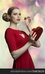 portrait of attractive young girl with pretty hair-style and make-up wearing red dress and taking in the hand her valentine gift, one box of chocolates