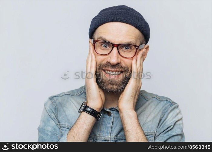 Portrait of attractive young man with thick beard and mustache looks with delighted expression, has white perfect teeth, poses in studio against blank copy space. People and positive emotions