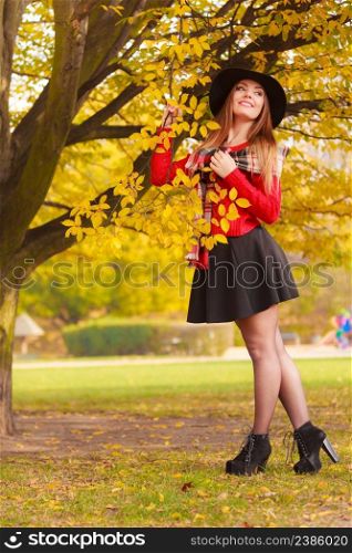 Portrait of attractive young smiling woman holding branch with leaves in autumnal park. Fashionable girl wearing red sweater scarf and black hat skirt high heels.. Beautiful woman in autumn park
